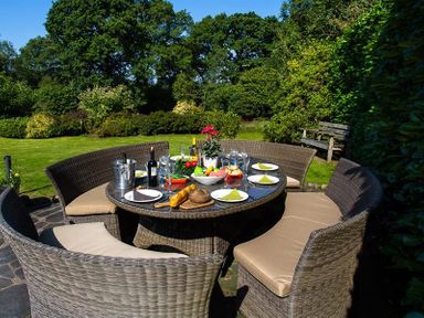An outdoor dining table with plates glasses bottles and bread surrounded by wicker chairs in a garden at Fair Rigg Old Farm in Cartmel