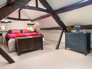 A bedroom with exposed wooden beams a bed with pink pillows and blanket a dark wooden chest and a black dresser with flowers at Fair Rigg Old Farm in Cartmel