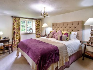 A bedroom with a bed with purple and beige bedding and floral curtains at Fair Rigg Old Farm in Cartmel