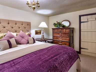 A bedroom with a bed with purple and beige bedding a wooden dresser with a mirror and a lamp on a round table at Fair Rigg Old Farm in Cartmel