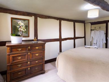 A bedroom with a wooden chest of drawers a plant on the chest a bed with beige bedding and two white robes hanging on the wall at Fair Rigg Old Farm in Cartmel