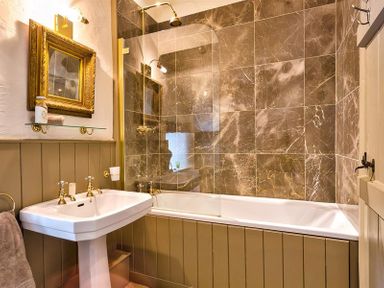 A bathroom with a white pedestal sink a bathtub with brown marble wall tiles and a gold framed mirror at Fair Rigg Old Farm in Cartmel