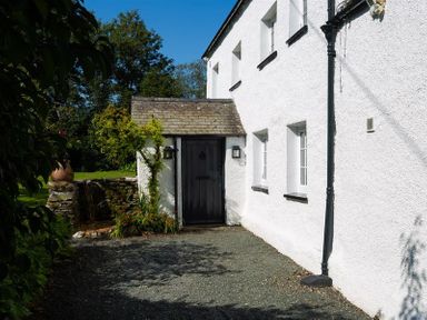 A white house exterior with a black door and small windows next to a garden at Fair Rigg Old Farm in Cartmel
