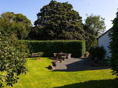 An outdoor garden with green grass trees a hedge a seating area and a white building at Fair Rigg Old Farm in Cartmel