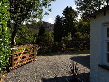 An outdoor gravel area with a wooden gate stone walls trees and a mountain in the background at Fair Rigg Old Farm in Cartmel