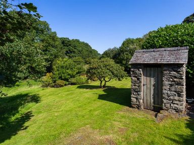 A grassy garden area with trees bushes and a small stone shed with a wooden door at Fair Rigg Old Farm in Cartmel