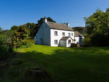 A white house with a slate roof surrounded by green lawn and trees at Fair Rigg Old Farm in Cartmel