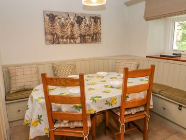 A dining area with a table covered by a lemon patterned cloth surrounded by wooden chairs and corner bench seating with cushions and a sheep painting on the wall at Low Cartmell Fold in Crosthwaite