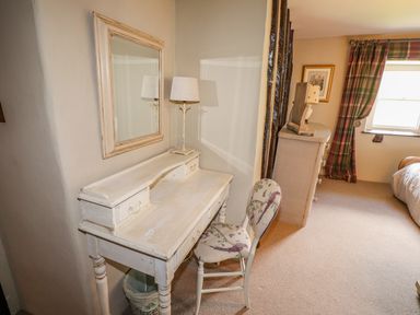 A bedroom corner with a white wooden desk and chair under a mirror next to a chest of drawers and a window with plaid curtains at Low Cartmell Fold in Crosthwaite