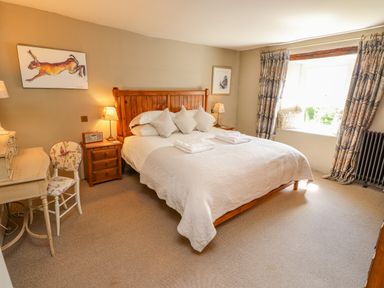 A bedroom with a wooden bed, bedside tables with lamps, a desk with a chair, and a window with patterned curtains at Low Cartmell Fold in Crosthwaite