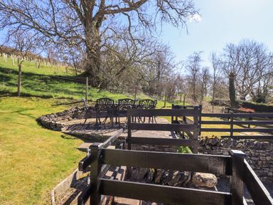 An outdoor patio with metal chairs and table near a grassy hill and trees at Low Cartmell Fold in Crosthwaite