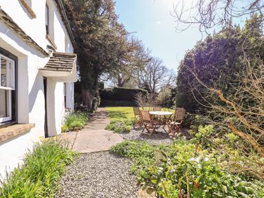 An outdoor garden area with a white house wall on the left wooden patio table and chairs on gravel and grass lawn with trees and shrubs at Low Cartmell Fold in Crosthwaite
