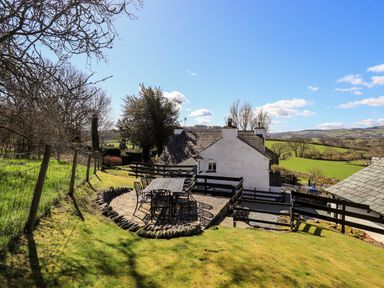 An outdoor patio with a metal table and chairs next to a white house with a tiled roof in a rural area at Low Cartmell Fold in Crosthwaite