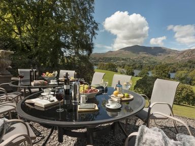 An outdoor patio with round glass tables set with wine glasses cups books fruit and bottles with chairs around overlooking hills and a lake at Tent Lodge in Coniston