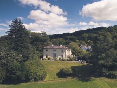 A house with a large lawn surrounded by trees under a partly cloudy sky at Tent Lodge in Coniston