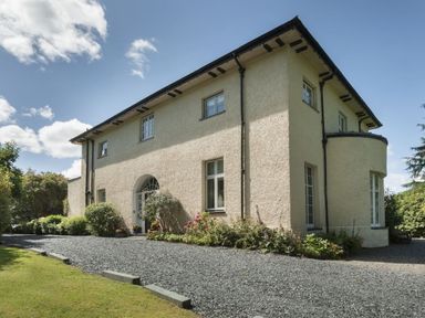A two-story beige stucco house with a gravel driveway and garden plants at Tent Lodge in Coniston