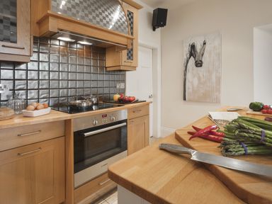 A kitchen with wooden cabinets, a built-in oven, a stovetop with pots, vegetables on the counter, and a horse painting on the wall at Tent Lodge in Coniston