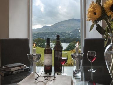 A table with two bottles of wine two lit candles two glasses of red wine and an open book with glasses by a window overlooking hills and a lake at Tent Lodge Coniston