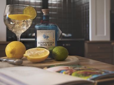 A kitchen countertop with a bottle of gin lemons limes a glass with lemon and lime slices and an open book