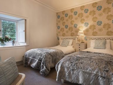 A bedroom with two single beds covered with gray fur blankets a nightstand with a lamp between the beds and a window with flowers on the sill at Tent Lodge in Coniston