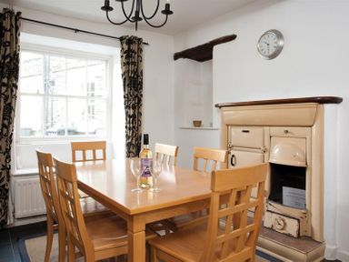 A dining room with a wooden table and chairs a bottle of wine three wine glasses a window with patterned curtains a wall clock and a cream colored stove at Pollys Cottage in Langdale