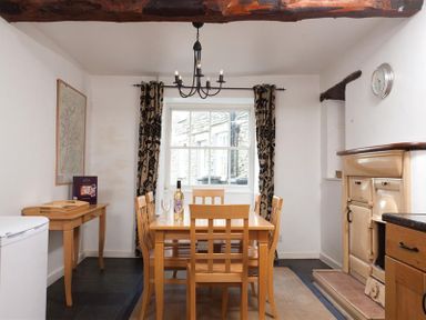 A dining room with a wooden table and six chairs a window with patterned curtains a wall clock a beige stove and a wooden side table at Pollys Cottage in Langdale