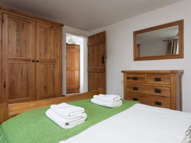 A bedroom with a wooden bed and green blanket with folded towels on it a wooden wardrobe a dresser with mirror and an open wooden door at Pollys Cottage in Langdale