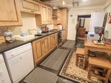 A kitchen with wooden cabinets a dishwasher a sink a gas stove a refrigerator a wooden dining table with two stools and striped rugs on the floor at Springwell Cottage Ambleside