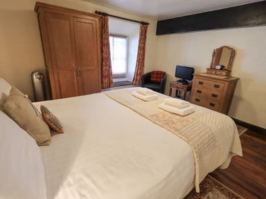 A bedroom with a bed with white linens two folded towels a wooden wardrobe a small television on a wooden dresser a black chair near a window with curtains at Springwell Cottage in Ambleside