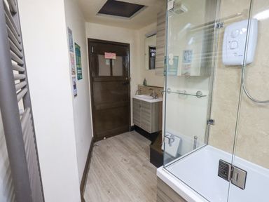 A narrow bathroom with a wooden door a sink with cabinet a bathtub with glass shower screen and a wall-mounted shower unit at Springwell Cottage in Ambleside