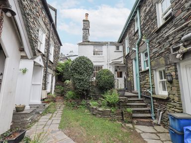 A stone cottage courtyard with paved path, shrubs, stone steps, and adjacent stone buildings at Springwell Cottage in Ambleside