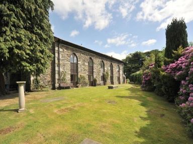A grassy garden with a stone building with arched windows and flower bushes at Candleberry Cottage in Ambleside