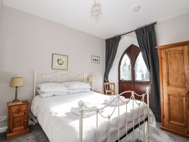 A bedroom with a white metal bed frame bedside tables lamps a wooden wardrobe and arched windows with dark curtains at Candleberry Cottage in Ambleside