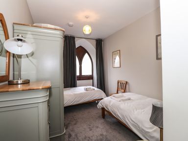 A bedroom with two single beds a wooden chair a wardrobe a lamp and an arched window at Candleberry Cottage in Ambleside