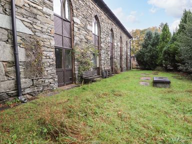 A stone building with arched windows and wooden doors next to a grassy area with benches and trees at Candleberry Cottage in Ambleside