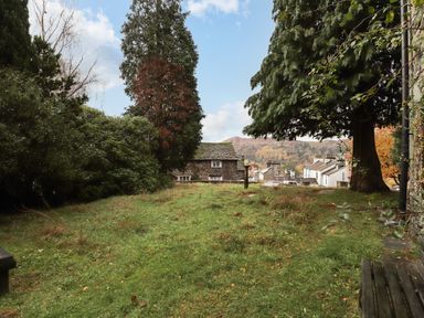 A grassy backyard with large trees and stone houses in the background at Candleberry Cottage in Ambleside
