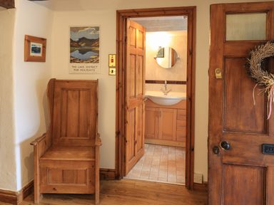 An entryway with a wooden chair a framed picture a door with a wreath and a view into a bathroom with a wooden cabinet and round mirror at Gale Lodge Cottage in Ambleside