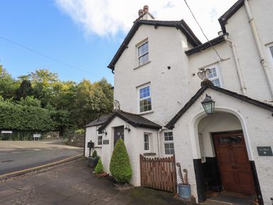 The exterior of a white cottage with a wooden door a small gate potted plants and signage on a paved street at Gale Lodge Cottage in Ambleside