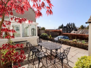 A patio with metal table and chairs surrounded by plants and a conservatory at Gale Lodge Cottage in Ambleside
