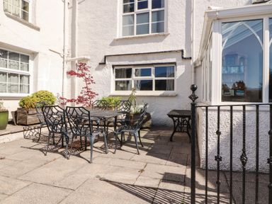 A patio with a metal table and six chairs next to a white building with windows and potted plants at Gale Lodge Cottage in Ambleside