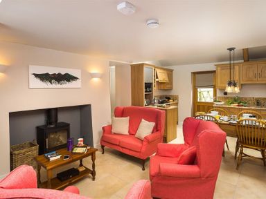 A living room with red upholstered armchairs and a sofa around a wood burning stove with a wooden coffee table and a dining area with wooden chairs at Barn Cottage in Bassenthwaite near Keswick