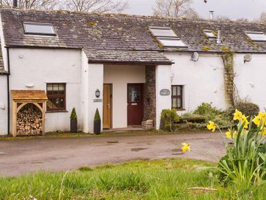 Exterior view of a cottage with stacked firewood and daffodil flowers in front at Barn Cottage Bassenthwaite near Keswick