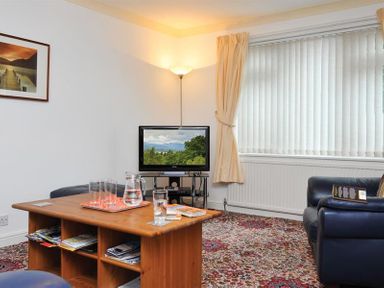 A living room with a wooden coffee table, glasses, a television on a stand, a floor lamp, a leather sofa, and a window with vertical blinds at Mosslea in Bowness