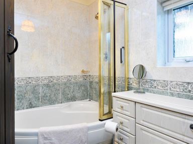 A bathroom with a corner bathtub with glass shower screen a white vanity unit with drawers and a small round mirror at Mosslea in Bowness