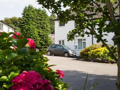 A street view with a grey car parked near a white house with bushes and pink flowers in the foreground at Mosslea in Bowness