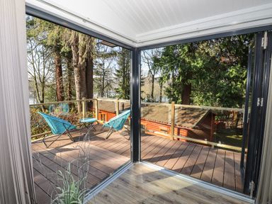 A balcony with two blue chairs and a small table overlooking trees and wooden buildings at Windermere Lodge in Bowness-On-Windermere