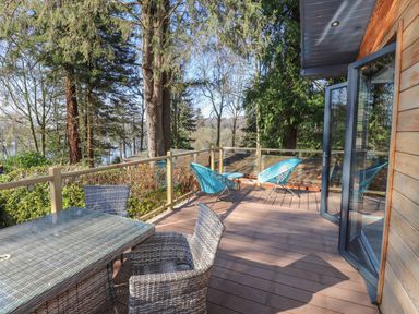 A wooden deck with a glass top table with wicker chairs and two blue chairs near a glass door surrounded by trees at Windermere Lodge in Bowness-On-Windermere