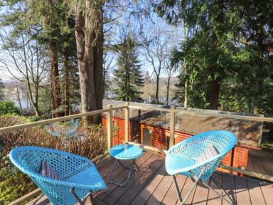 A balcony with two blue chairs and a small table overlooking trees and a wooden building at Windermere Lodge in Bowness-On-Windermere