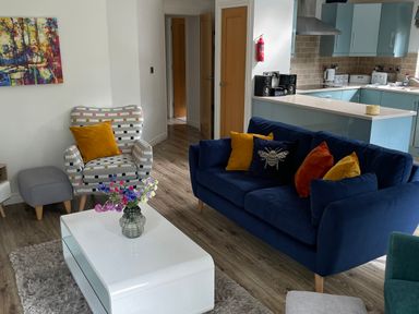 A living room with a blue sofa and colorful cushions a patterned armchair a white coffee table with flowers and an open kitchen at Windermere Lodge in Bowness-On-Windermere