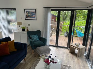 A living room with a blue sofa and armchair a white cabinet and glass doors leading to a wooden deck at Windermere Lodge in Bowness-On-Windermere